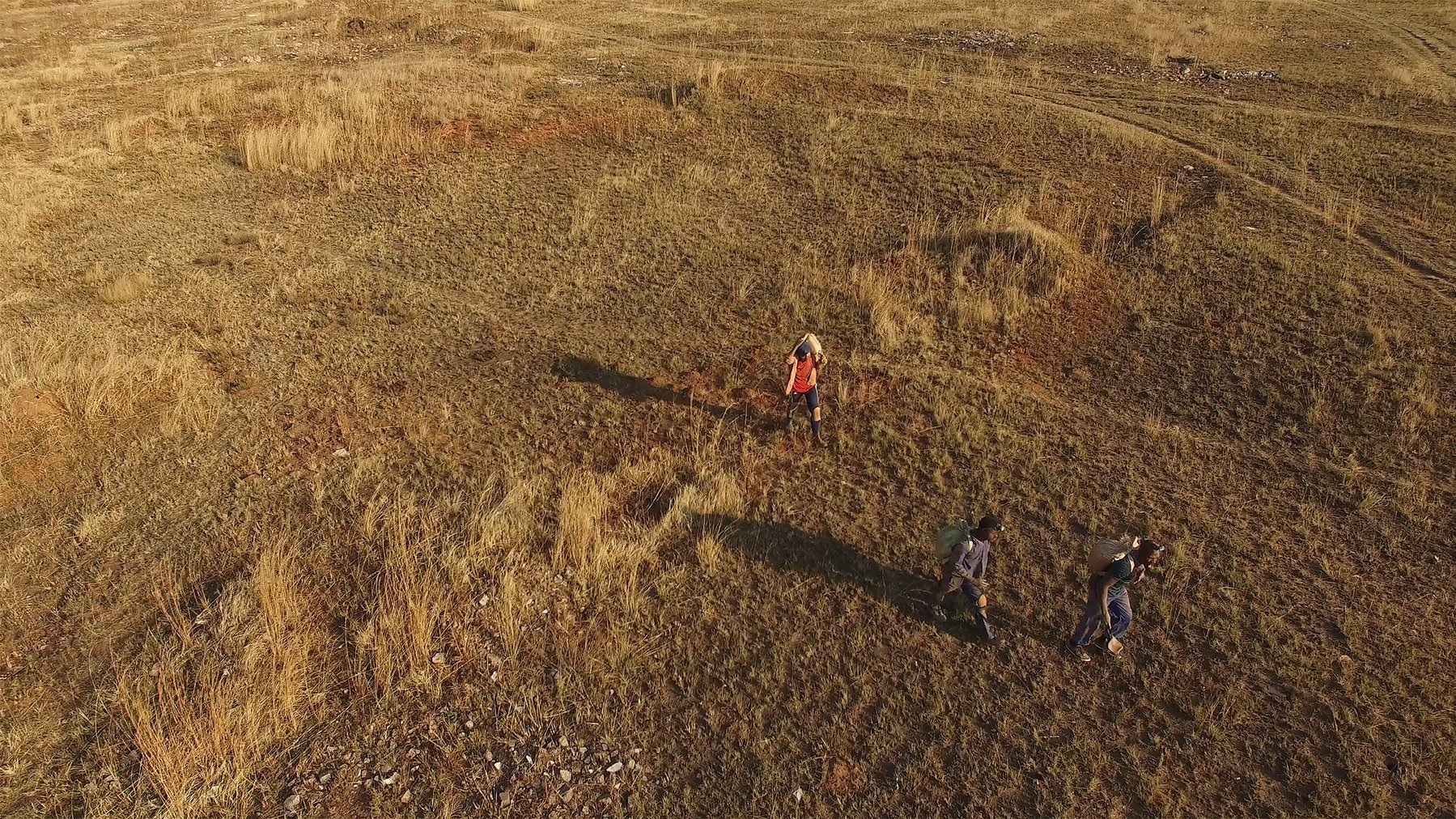 Dans cette image, on voit un paysage vaste et ouvert, couvert d'herbe sèche et de petites touffes de plantes. Le sol est légèrement accidenté, avec des zones de terre claire et quelques pierres éparpillées. Trois personnes marchent dans ce terrain. L'une d'elles porte un vêtement rouge, tandis que les autres sont habillées de couleurs plus sombres. Leur ombre s'étend sur le sol, suggérant que le soleil est assez bas dans le ciel. L'atmosphère est calme et naturelle, évoquant une promenade en pleine nature.