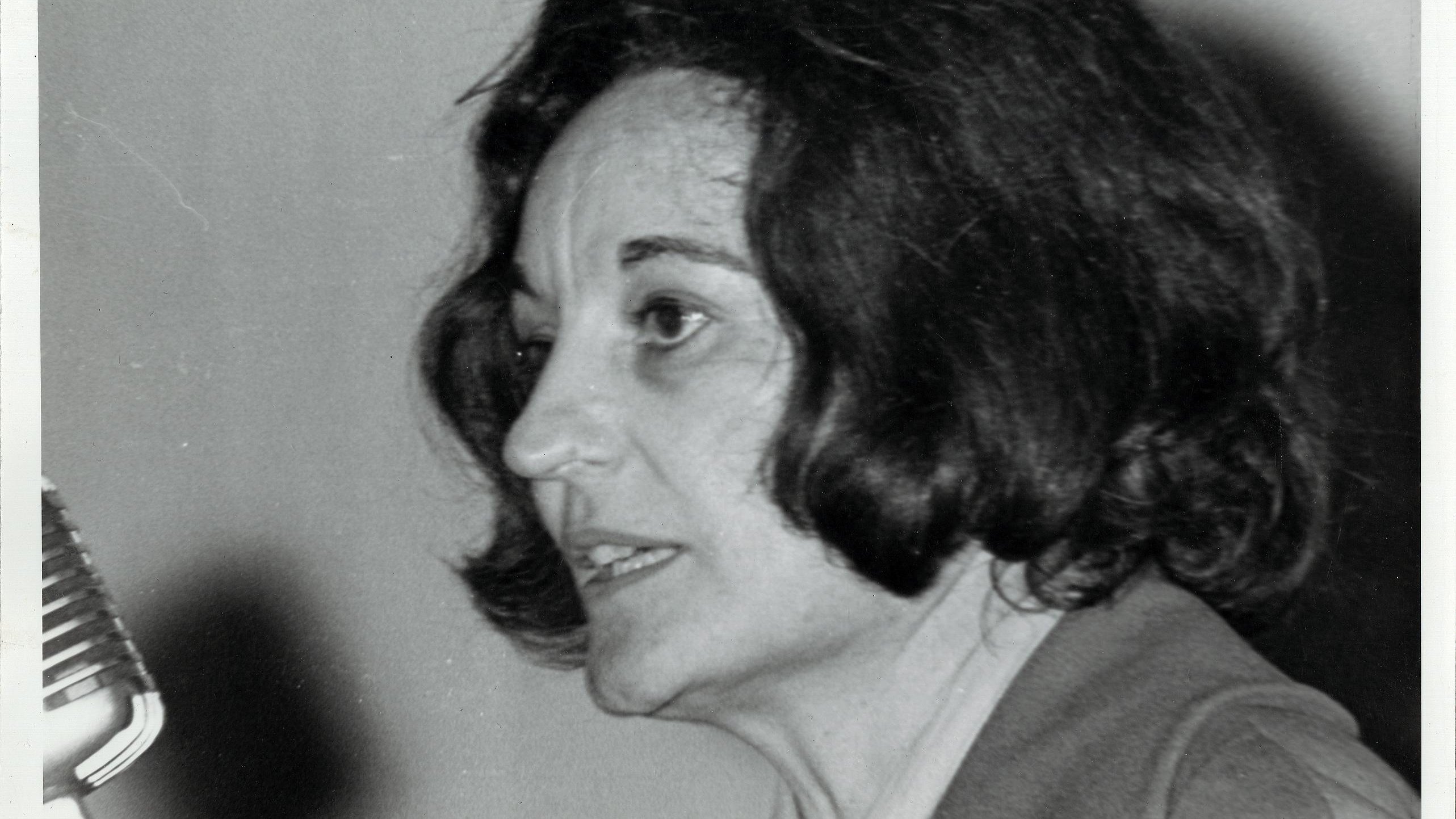 L'image montre une femme avec des cheveux foncés, mi-longs et légèrement ondulés. Elle est en profil, parlant près d'un microphone. Son expression est concentrée, et on peut deviner une certaine passion dans son discours. Elle porte une veste simple, et l'arrière-plan est flou, ce qui met l'accent sur son visage et son expression. L'éclairage est doux, soulignant les traits de son visage.