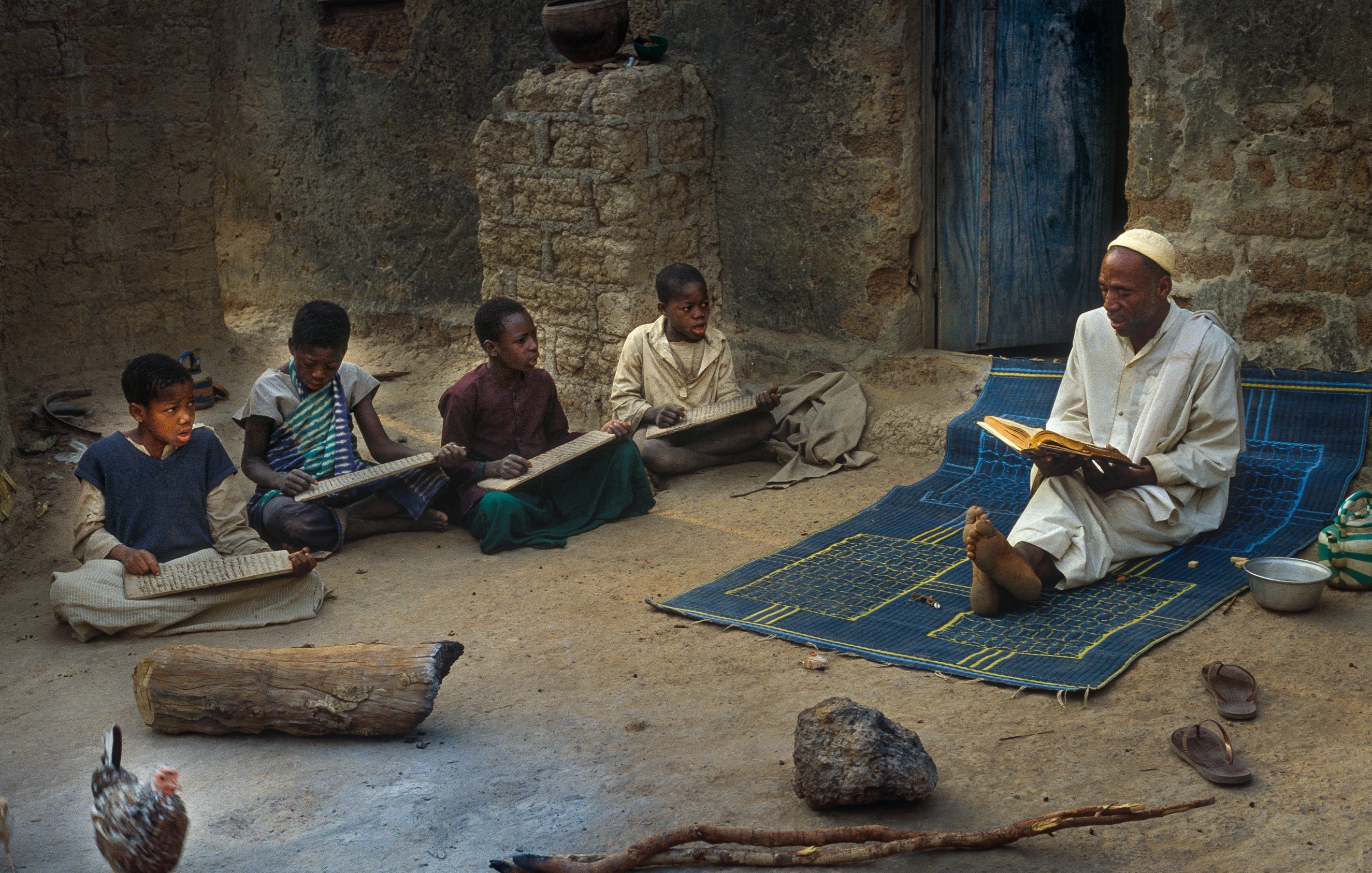 L'image montre un enseignant assis sur un tapis à l'extérieur d'une maison en terre. Il lit un livre avec attention, tandis que plusieurs enfants sont rassemblés autour de lui, concentrés sur des planches ou des livres qu'ils tiennent. L'ambiance est calme et studieuse. On peut aussi voir un poulet se déplacer au sol, ajoutant une touche de vie rurale à la scène. Les murs de la maison sont simples, et la lumière naturelle éclaire l'espace, créant une atmosphère chaleureuse et conviviale.