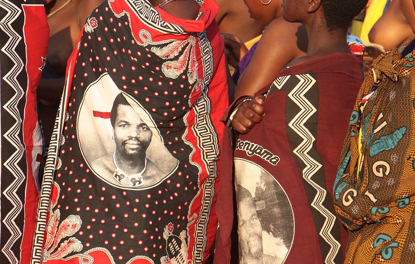 Dans cette image, on voit un groupe de femmes habillées de façon traditionnelle, revêtues de vêtements colorés et ornés de motifs riches. Certaines portent des châles qui affichent des portraits d'un homme, probablement une figure importante de leur culture. Les femmes se tiennent proches les unes des autres, exprimant un esprit de solidarité. L'atmosphère est vibrante, suggérant une célébration ou un événement culturel, avec des couleurs vives qui évoquent une culture riche et dynamique. Les sons environnants pourraient comprendre des musiques folkloriques et des rires, ajoutant à l'ambiance festive.