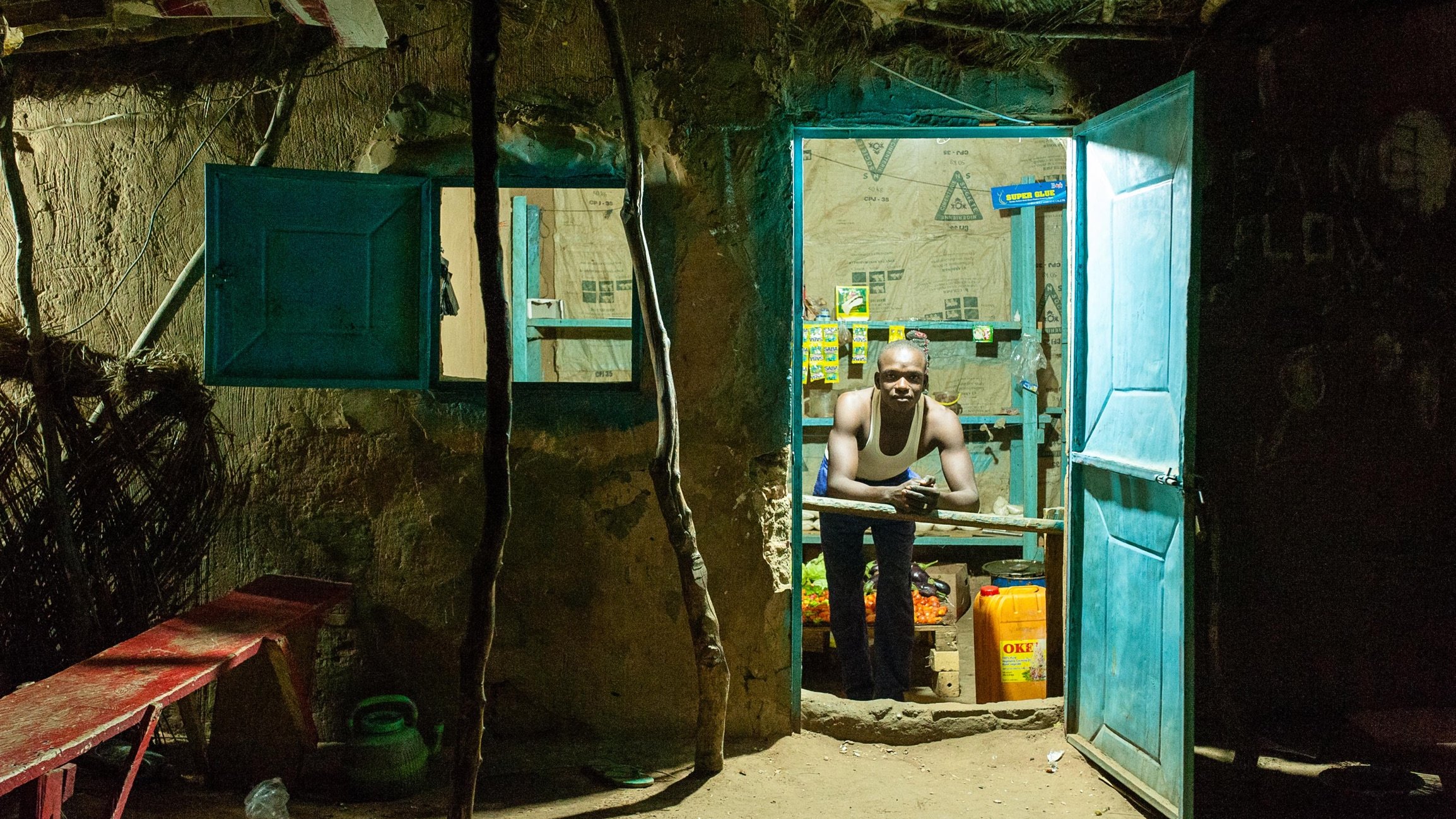 L'image montre l'entrée d'une petite boutique, éclairée dans l'obscurité. La porte est ouverte, révélant un intérieur modeste. On aperçoit un homme qui se tient derrière un comptoir, son regard tourné vers l'extérieur. Il porte un débardeur et semble attentif, probablement en attente de clients. Les murs sont en terre, et des papiers et affiches sont accrochés, ajoutant une touche de couleur à l'environnement. À l'avant, un banc en bois rouge est installé, et quelques bouteilles et contenants sont visibles à l'intérieur de la boutique. L'ambiance est chaleureuse malgré le contexte rural et simple.