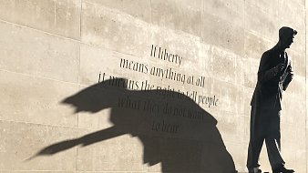 L'image représente une statue d'un homme debout, probablement un auteur ou un penseur, sur un socle. La statue est en bronze, et son ombre projette une forme dynamique sur le mur derrière elle. Ce mur est en pierre lisse, et il porte une citation célèbre qui commence par « If liberty means anything at all... », reflétant des thèmes de liberté et d'expression. L'ensemble crée une atmosphère réfléchie et inspirante, incitant à considérer la profondeur des mots inscrits ainsi que le rôle de la statue.