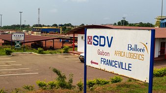 L'image montre un panneau indicatif dans une zone logistique. Ce panneau, situé devant un bâtiment, indique la direction vers Franceville, une ville du Gabon. On peut apercevoir le logo de SDV, ainsi que celui de Bolloré Africa Logistics. Le paysage environnant est assez verdoyant, avec plusieurs arbres et buissons. Au loin, on distingue une infrastructure de transport, possiblement liée à la logistique. L'atmosphère est calme, reflétant un lieu dédié aux activités commerciales et de logistique.