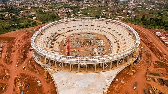 L'image montre un stade en cours de construction, vu d'en haut. Le stade a une forme ovale et un grand espace intérieur qui est encore vide, avec des structures en cours d'installation. Autour du stade, on peut voir un terrain rouge, probablement une terre fouillée pour la construction. À l'extérieur de la structure, il y a des machines de construction et des matériaux éparpillés. À l'arrière-plan, des maisons et des collines sont visibles, ce qui donne une idée de l'environnement urbain et naturel qui entoure le stade. L'ensemble de la scène évoque une ambition de développement et de modernisation.