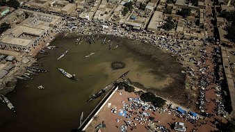Imaginez un paysage vu du ciel. Au centre, un plan d'eau sombre qui semble être une lagune ou un port. Les bords de l'eau sont bordés de bateaux, certains amarrés, d'autres flottant paisiblement. Autour de cette lagune, il y a des habitations, des maisons en terre ou en tuiles, formant un petit village. Sur les rives, des gens sont rassemblés, certains assis par terre, d'autres se déplaçant avec des marchandises. Des tentes et des parasols sont disposés, suggérant un marché vivant où l'on troque et vend des produits. L'air est probablement rempli de sons, de cris et d'odeurs de nourriture, de poissons et de marchandises variées, donnant l'impression d'une scène animée et prospère. Les couleurs sont chaudes, évoquant un climat ensoleillé. Ce tableau dégage une atmosphère d'activité et de communauté autour de l'eau.