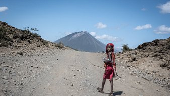 L'image montre un enfant marchant sur un chemin de terre. Il porte une robe rouge et un foulard, et il semble porter un petit récipient ou une bouteille dans ses mains. En arrière-plan, on aperçoit un volcan majestueux, dont le sommet est légèrement nuageux sous un ciel bleu clair parsemé de quelques nuages blancs. Le terrain autour est rocailleux et aride, avec quelques petits buissons éparpillés. L'ensemble de la scène dégage une atmosphère de nature sauvage et éloignée.