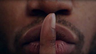 L'image présente un gros plan sur un visage, principalement la zone des lèvres et du nez. Un doigt est placé devant la bouche, formant le geste universel du « chut ». La peau est de teinte foncée, et les lèvres sont bien définies, affichant une légère brillance. Le regard de la personne est probablement dirigé vers l'extérieur de l'image, transmettant une impression de mystère ou de secret. L'arrière-plan est flou, ce qui met l'accent sur le geste et l'expression du visage.