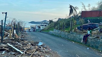 L'image présente une scène de destruction dans une rue. On voit des débris en bois éparpillés sur le sol, témoignant d'un événement dévastateur, peut-être une tempête ou un ouragan. Sur la gauche, il y a des maisons endommagées, certaines avec des toits manquants. En arrière-plan, on devine un paysage côtier avec une étendue d'eau qui reflète la lumière du ciel. À droite, une personne est assise sur un mur, habillée de vêtements colorés, semblant contempler les dégâts. La lumière du jour est douce, annonçant un crépuscule paisible, contrastant avec la morosité de la scène. Les palmiers, certains déracinés, ajoutent à l'impression de désolation.