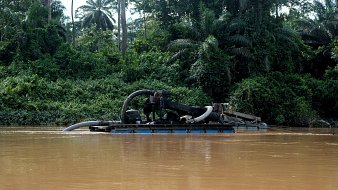 L'image montre un bateau sur une rivière aux eaux brunes. Ce bateau est équipé d'un grand tuyau qui semble aspirer de la matière de l'eau. À l'arrière-plan, on peut voir une végétation dense, composée de grandes plantes et d'arbres tropicaux, qui créent une ambiance naturelle et verdoyante. La scène évoque une activité liée à l'extraction de ressources, probablement de l'or ou des minéraux, dans un environnement de jungle.