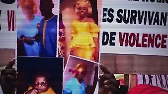 L'image montre une scène de manifestation où des participants tiennent des pancartes. Les pancartes contiennent des messages forts sur le soutien aux survivantes de la violence. Certaines affichent des photos, peut-être de personnes ou d'enfants, ainsi qu'une poupée, ce qui suggère une connexion émotionnelle ou un symbole de douceur au milieu d'un discours poignant. L'ambiance est intacte, avec des slogans qui crient l'injustice et l'urgence de protéger ceux qui ont souffert.