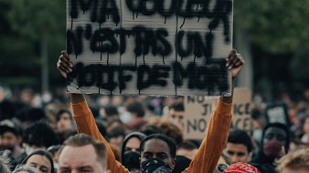 L'image montre une grande foule rassemblée pour une manifestation. Parmi les manifestants, une personne se distingue en tenant une pancarte sur laquelle est écrit "MA COULEUR N'EST PAS UN CRIME". Cette personne porte un masque, comme beaucoup d'autres dans la foule. Les visages autour sont sérieux et déterminés, reflétant un sentiment de solidarité et de revendication. On peut sentir l'intensité de l'événement grâce à l'énergie collective. En arrière-plan, des arbres indiquent un cadre naturel, ajoutant à l'atmosphère de mobilisation pacifique.