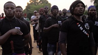 L'image montre un groupe de jeunes hommes debout en formation. Ils portent des t-shirts noirs et semblent participer à une activité collective, probablement un chant ou un cri de ralliement. Certains d'entre eux tiennent des bâtons ou des armes. En arrière-plan, on aperçoit d'autres personnes rassemblées, avec des arbres et un paysage rural. L'atmosphère semble être à la fois solennelle et engagée. La lumière du soir crée une ambiance particulière.
