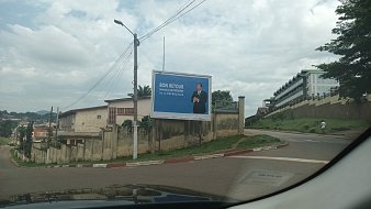 L'image montre une grande affiche publicitaire installée sur un mur à l'extérieur. Sur cette affiche, un homme, probablement un homme politique, se tient debout avec un sourire, vêtu d'un costume. Le fond est bleu, ce qui attire l'attention. En haut de l'affiche, on peut lire le texte "BON RETOUR", suivi d'un message qui semble faire référence à un événement ou une situation spécifique. En dessous, s'étend un paysage urbain avec des bâtiments, des routes et des arbres, et l'image suggère un périphérique ou une route en pente. Le ciel est partiellement nuageux, ajoutant une ambiance calme à la scène.