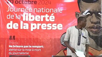 L'image montre un événement lié à la Journée nationale de la liberté de la presse, qui se déroule le 20 octobre 2024. En arrière-plan, un grand panneau rouge présente le thème de la journée : "Ne brisons pas le rempart : alerte sur la mise à mort du journalisme". Au premier plan, un homme debout s'apprête à s'adresser à l'audience. Il porte une chemise à rayures noires et blanches et se tient près d'une table équipée de plusieurs microphones et de bouteilles d'eau. L'ambiance est sérieuse et engageante, soulignant l'importance de la liberté d'expression.