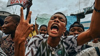 L'image montre une scène de manifestation animée. Au premier plan, un jeune homme crie avec passion, levant les deux doigts en signe de victoire ou de protestation. Son visage exprime une forte intensité émotionnelle, avec une bouche ouverte et des yeux déterminés. Il porte un maillot à motifs verts et blancs, représentant probablement les couleurs de son pays. Derrière lui, un groupe de personnes brandit des pancartes avec des slogans, illustrant un sentiment de mobilisation et de solidarité. L'ambiance est vibrante, évoquant une lutte collective pour des causes qui leur tiennent à cœur, sous un ciel nuageux.