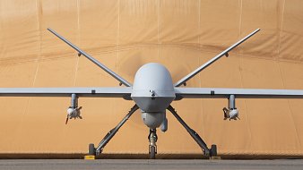 L'image montre un drone militaire de type MQ-9 Reaper. Il est positionné de face, avec le corps fuselé et l'aile légèrement étendue. À chaque côté du fuselage, on peut voir des réservoirs ou des munitions. En arrière-plan, il y a une toile beige qui semble un hangar ou une protection, lui donnant un aspect de préparation à un vol. Le sol est en béton, créant un environnement d'aérodrome. L'ensemble est impressionnant, évoquant la technologie aéronautique moderne.