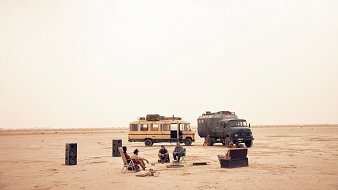 L'image montre un paysage désertique, vaste et aride, baigné par une lumière douce. Au centre, un groupe de personnes est assis autour d'un feu de camp. Ils semblent se détendre et passer un bon moment ensemble. À proximité, deux véhicules distincts attirent l'attention : l'un est un camping-car jaune, l'autre un véhicule utilitaire de couleur sombre. Des haut-parleurs ont été disposés autour du groupe, évoquant une ambiance festive. Le sol est sablonneux, et l'atmosphère semble calme, presque magique, sous un ciel d'une teinte uniforme.