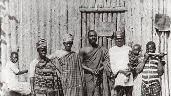 L'image montre un groupe de six personnes se tenant debout devant une structure en bois, qui semble être une maison ou un abri, avec un décor rustique. Au fond, on distingue un homme assis qui fait face à la scène principale. Les membres du groupe portent des vêtements traditionnels typiques, avec des motifs variés, affichant une richesse culturelle. L'un d'eux tient un enfant, tandis qu'un autre est équipé d'un bâton, suggérant une certaine autorité ou dignité. L'atmosphère de l'image dégage un sentiment de communauté et de convivialité.