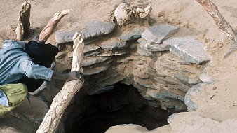 L'image montre un trou dans le sol, probablement un puits, entouré de pierres disposées de manière circulaire. À côté du trou, on aperçoit une personne, vêtue d'une chemise et d'un pantalon longs, qui semble s'approcher ou manipuler un morceau de bois. Le terrain est sablonneux, et des débris de bois sont visibles autour, suggérant une utilisation régulière de cet endroit. L'atmosphère semble aride, reflétant un environnement désertique.