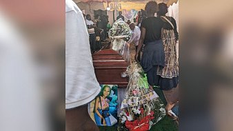 L'image montre un moment poignant d'un enterrement. Au centre, un cercueil en bois brun, sur lequel sont déposés des bouquets de fleurs. À côté du cercueil, une grande photo d'une femme est accrochée, entourée de décorations florales et de bandes de tissu. Des personnes, visiblement en deuil, se rassemblent autour, certaines portant des vêtements sombres. L'ambiance est sombre mais respectueuse, marquée par la tristesse et l'hommage à la personne décédée. L'arrière-plan est flou, mais on peut deviner une tente qui abrite cette cérémonie. L'ensemble évoque des sentiments de perte et de commémoration.