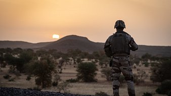 La scène se déroule dans un paysage désertique au coucher du soleil. Au premier plan, un soldat en uniforme camouflage se tient debout, tourné vers l'horizon. Sa silhouette est mise en valeur par la lumière dorée du soleil qui descend lentement derrière une colline arrondie. Le terrain est rocailleux et parsemé de petites végétations et arbres, typiques des paysages arides. L'ambiance est calme, presque contemplative, ajoutant une dimension de réflexion à la présence du soldat, probablement en mission. Les couleurs chaudes du ciel se mélangent à la beauté naturelle du paysage, créant une atmosphère à la fois sereine et intense.