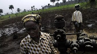 L'image montre un paysage rural où se trouvent des plantations de palmiers en arrière-plan. Au premier plan, une femme d'âge mûr, vêtue d'une blouse rayée et portant un foulard coloré, se tient près d'une infrastructure de forage pétrolier. Son expression est sérieuse et préoccupée. À l'arrière-plan, une autre personne, également en tenue décontractée, semble s'occuper de l'équipement. Le sol est noirci, suggérant une dégradation environnementale. L'atmosphère générale évoque une lutte pour préserver leur lien avec la terre face aux impacts de l'exploitation pétrolière.
