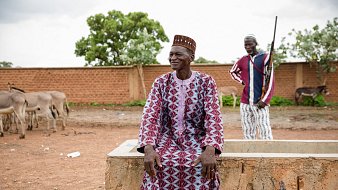 L'image montre un homme assis sur un mur en béton dans un environnement rural. Il porte une tenue traditionnelle, ornée de motifs géométriques dans des tons de rouge et blanc, et un chapeau conique typique. Son expression est détendue et souriante, suggérant une ambiance joyeuse. À l'arrière-plan, un deuxième homme se tient debout, avec un fusil à la main, veillant sur des ânes qui paissent paisiblement. La scène évoque une vie simple et traditionnelle, ancrée dans la nature. Le ciel est nuageux, ajoutant une légère atmosphère de sérénité au paysage.