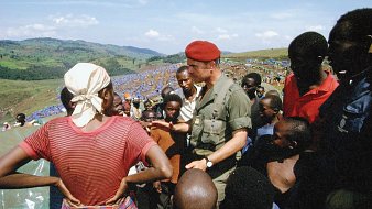 L'image montre un groupe de personnes dans un paysage ouvert et verdoyant. Au centre, un homme en uniforme militaire, portant un béret rouge, discute avec une femme qui porte un foulard et une blouse, le regard tourné vers lui. Elle semble attentive aux explications qu'il donne. Autour d'eux, plusieurs enfants et adultes, principalement d'origine africaine, écoutent avec intérêt. Les visages sont expressifs, certains avec des regards curieux. À l'arrière-plan, on aperçoit une vaste étendue de tentes bleues, indiquant probablement un camp de réfugiés ou de soutien. Le ciel est clair, avec quelques nuages, et les collines verdoyantes offrent une vue panoramique sur la scène. L'atmosphère dégage un sentiment de communauté et d'engagement.