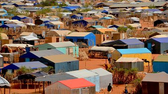 L'image montre un vaste camp de tentes et de logements temporaires disposés de manière dense sur un terrain désertique. Les structures sont recouvertes de toits de différentes couleurs, principalement des teintes de bleu, de rouge et de gris. Le sol est sablonneux, et quelques arbres ou buissons apparaissent ici et là, apportant un peu de verdure. En bas de l'image, on peut apercevoir des personnes se déplaçant entre les tentes, évoquant une activité quotidienne dans ce milieu. L'ensemble dégage une impression d'organisation dans la diversité des abris, tandis que le paysage aride souligne les conditions de vie dans ce camp.