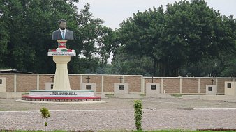 Cette image montre une statue en l'honneur d'une personnalité importante, probablement un leader ou un héros national. La statue est placée sur un socle élevé et représente un homme, vêtu d'un costume. En arrière-plan, on peut voir un mur avec plusieurs croix, qui semblent être des stèles commémoratives. Le site est entouré de verdure, avec des arbres touffus qui apportent une atmosphère paisible. Le sol est en pavé, et des plantes décoratives sont présentes, ajoutant une touche de nature à cet espace de mémoire.