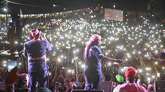 L'image montre une scène d'un concert ou d'un rassemblement en plein air, où un grand nombre de personnes est rassemblé devant un podium. Au premier plan, deux artistes sont sur scène, l'un chantant et l'autre en mouvement, tous deux vêtus de vêtements décontractés, avec des chapeaux et des bandanas. Derrière eux, une mer de spectateurs se tient en arrière-plan, illuminée par d'innombrables lumières de téléphones portables levés, créant un effet scintillant dans la nuit. L'atmosphère est électrisante, avec des couleurs vives provenant des vêtements des membres du public et des affiches. Des banderoles et des drapeaux sont également visibles, ajoutant à l'ambiance festive et engagée de l'événement.