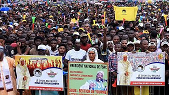 L'image montre une grande foule rassemblée pour un événement ou un meeting public. Les gens se tiennent serrés les uns contre les autres, créant une mer de visages et de vêtements colorés. De nombreux participants tiennent des banderoles avec des messages politiques, notamment le slogan "Le choix du peuple pour le changement." On peut également apercevoir des drapeaux flottants, évoquant une atmosphère de mobilisation et d'engagement. L'arrière-plan suggère une ambiance urbaine, avec des bâtiments visibles au loin, tandis que le ciel pourrait être nuageux ou couvert. Ce rassemblement dégage un sentiment d'énergie collective, d'espoir et de désir de changement.