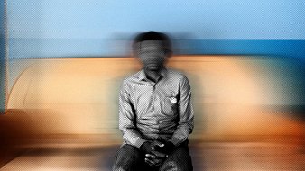 Cette image montre une personne assise sur un canapé. Le fond est flou, ce qui donne une impression de mouvement. La personne porte une chemise à rayures et a les mains posées sur ses genoux. L'atmosphère de l'image est douce grâce aux couleurs chaudes du canapé et aux nuances de bleu derrière. L'ensemble crée une sensation de calme, mais le flou ajoute une dimension de mystère.