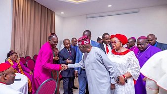 L'image montre un groupe de personnes rassemblées dans une salle, probablement lors d'un événement formel. Au centre, un homme en tenue traditionnelle serre la main d'un autre homme, qui porte une chemise claire. Les autres participants, habillés de manière variée, se tiennent autour d'eux. Certaines femmes portent des vêtements colorés et des accessoires élégants. L'ambiance semble conviviale et respectueuse, avec des sourires et des gestes amicaux. Les murs de la salle sont de couleur claire, et il y a des chaises rouges disposées autour de tables.