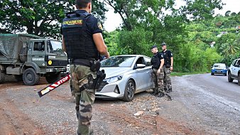L'image montre un contrôle routier en plein air. Au premier plan, un gendarme en uniforme, portant un gilet par balles noir avec l'inscription "Gendarmerie", observe une voiture argentée qui est arrêtée sur le côté de la route. La voiture a un panneau "STOP" visible. Deux autres gendarmes, habillés de manière similaire, se tiennent près de la voiture, dialoguant probablement avec le conducteur. En arrière-plan, on aperçoit une camionnette militaire de la gendarmerie et des arbres verdoyants qui donnent une ambiance naturelle à la scène. Le sol est en terre, avec beaucoup de verdure autour, ce qui indique que le lieu est rural. L'atmosphère semble sérieuse, reflet du contrôle de sécurité en cours.