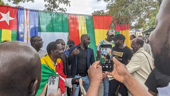 L'image montre un groupe de personnes réunies autour d'un fond coloré constitué de drapeaux. Les drapeaux affichés incluent ceux du Cameroun, de l'Union européenne, du Mali et de la République de Guinée. Des hommes portent des vêtements variés, certains drapés dans des drapeaux, ce qui suggère un événement festif ou culturel. Ils semblent en discussion animée, souriant et échangeant des plaisanteries. Au premier plan, un participant tient un téléphone portable, probablement pour capturer le moment. L'atmosphère semble conviviale et pleine d'énergie.