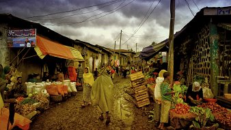 Cette image dépeint une scène de marché dans une rue étroite et animée. Le sol est boueux, probablement à cause de la pluie récente. Des étals colorés, couverts de bâches orange et bleues, sont remplis de fruits et légumes frais. Des vendeurs, portant des vêtements traditionnels et des capuchons pour se protéger de l'humidité, interagissent avec les clients. On peut ressentir l'effervescence du marché, avec des gens qui marchent, négocient et échangent des sourires. Au fond, le ciel est nuageux, ajoutant une atmosphère grise à cette scène vivante. Ce mélange de sons, d'odeurs et d'activités crée une ambiance dynamique et chaleureuse, typique des marchés locaux.