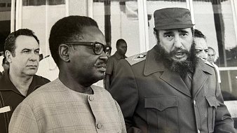 Cette image en noir et blanc montre trois hommes en discussion. Au premier plan, deux hommes sont particulièrement visibles. À droite, un homme avec une barbe fournie et un uniforme militaire, caractéristique de Fidel Castro, dialogue avec un homme portant des lunettes et un costume clair. Ce dernier semble attentif à la conversation. En arrière-plan, un troisième homme observe la scène. L'environnement est urbain, avec des bâtiments à vitre en arrière-plan. L'atmosphère paraît sérieuse et engagée, suggérant une rencontre diplomatique. Les expressions des visages laissent transparaître une certaine gravité.