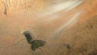 L'image montre une vaste étendue de paysage désertique, dominée par des teintes de beige et de marron clair. On aperçoit des stries blanches qui semblent indiquer des vents ou des mouvements de sable dans l'air. En bas de l'image, un petit plan d'eau apparaît, entouré de terres arides. Ce contraste entre la surface sèche du désert et l'eau brillante crée une scène à la fois aride et intrigante, témoignant des forces naturelles en jeu dans cet environnement désertique. Les textures et les couleurs évoquent une ambiance silencieuse et vaste, typique des paysages désertiques.