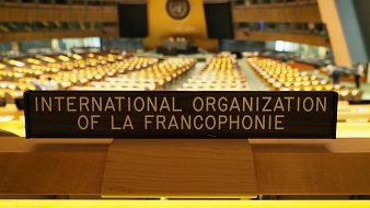 Cette image montre un intérieur d'une grande salle de conférence, probablement à l'ONU. Au premier plan, on voit un panneau en bois portant l'inscription "INTERNATIONAL ORGANIZATION OF LA FRANCOPHONIE". Derrière ce panneau, de nombreuses rangées de sièges sont disposées, chaque rangée étant équipée de petits dispositifs lumineux. La lumière ambiante de la salle crée une atmosphère chaleureuse et accueillante. Les couleurs varient entre des teintes dorées des dispositifs lumineux et les éléments architecturaux modernes du bâtiment. On peut sentir un sentiment d'importance et de sérieux dans cet espace, dédié à des discussions diplomatiques et internationales.