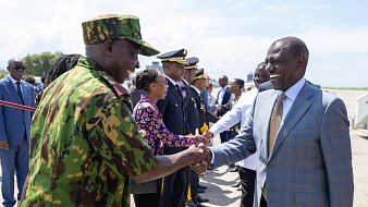 L'image montre une scène de bienvenue officielle à l'aéroport. Au premier plan, deux hommes se serrent la main, exprimant une atmosphère de camaraderie et de respect. L'un des hommes est en uniforme militaire avec un motif camouflage, tandis que l'autre porte un costume élégant avec une cravate. Autour d'eux, un groupe de personnalités, dont des membres des forces de sécurité et des fonctionnaires en civil, se tient en ligne, attendant leur tour pour saluer. En arrière-plan, le ciel est clair et ensoleillé, créant une ambiance positive. L'ensemble de la scène respire la solennité et l'importance d'un événement officiel.