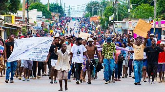 L'image montre une foule nombreuse marchant dans une rue animée. Les gens sont regroupés, certains tiennent des banderoles ou des pancartes. L'ambiance semble dynamique, avec beaucoup de mouvements et des échanges d'expressions entre les participants. On peut également ressentir une diversité dans les vêtements, allant des tenues décontractées aux vêtements plus colorés. En arrière-plan, des boutiques et des bâtiments bordent la rue, tandis que des fils électriques sont suspendus au-dessus. Les voix et les chants des manifestants semblent animer l'espace, créant une atmosphère de rassemblement et de mobilisation.