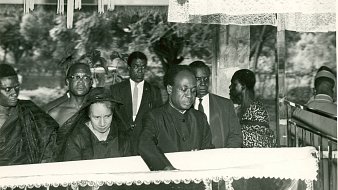 L'image présente une scène de recueillement, probablement lors d'un enterrement ou d'une commémoration. Au premier plan, un homme, habillé en noir, se penche vers un cercueil reposant sur une estrade, symbolisant le respect et la tristesse. À ses côtés se trouve une femme, également en noir, qui semble très émue. En arrière-plan, plusieurs personnes regardent la scène avec un air sérieux, toutes vêtues de manière formelle, ce qui souligne le caractère solennel de l'événement. L'atmosphère est empreinte de respect et de recueillement, avec une ambiance calme, interrompue seulement par les murmures des participants.