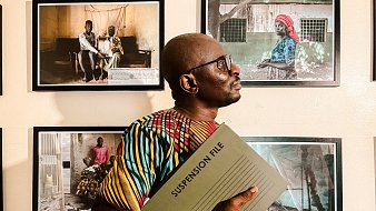 L'image montre un homme debout dans un espace d'exposition, entouré de plusieurs photographies accrochées au mur. Il porte une chemise colorée aux motifs vibrants, mêlant des teintes de jaune, rouge et vert. Dans ses bras, il tient un dossier intitulé "SUSPENSION FILE", ce qui suggère qu'il se prépare à discuter ou à présenter quelque chose d'important. L'homme se tourne légèrement sur le côté, regardant vers les photos, qui semblent raconter des histoires de vie, avec des portraits d'autres personnes. L'atmosphère de l'image est à la fois sérieuse et engagée, soulignant l'importance des histoires capturées dans ces œuvres visuelles.