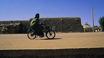 L'image montre un homme en train de rouler à bicyclette sur une route en terre. Il porte une tenue traditionnelle de couleur verte, qui contraste avec le décor. En arrière-plan, on peut apercevoir des bâtiments en adobe, typiques des régions désertiques, avec des murs de terre et des fenêtres. Le ciel est d'un bleu éclatant, sans nuages, et l'atmosphère générale est calme, suggérant une journée ensoleillée. La lumière du soleil éclaire la scène, mettant en valeur les textures du sol et des constructions.