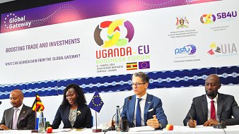L'image montre une scène d'un forum d'affaires intitulé "Uganda-EU Business Forum". Au premier plan, quatre personnes sont assises à une table, chacune ayant un micro devant elle. À gauche, un homme souriant semble représenter l'Ouganda. Ensuite, une femme, également souriante, prend la parole. Au centre, un homme, avec des lunettes et un air sérieux, est en train de s'exprimer. Enfin, à droite, un autre homme, avec une posture attentive, écoute. En arrière-plan, une banderole avec des logos et le titre de l'événement est accrochée. L'ambiance est professionnelle, et l'événement semble axé sur le renforcement des échanges et des investissements entre l'Uganda et l'Union européenne.