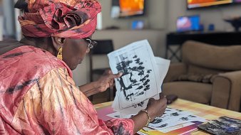 Dans cette image, une femme d'âge mûr, vêtue d'une magnifique robe rose et d'un foulard assorti, est assise sur un canapé dans un salon lumineux. Elle feuillette attentivement des photos en noir et blanc, toutes étalées devant elle sur une table basse recouverte d'une nappe à carreaux jaune et blanc. Le décor du salon est moderne, avec des télévisions visibles en arrière-plan, reflétant une ambiance chaleureuse et accueillante. La concentration de la femme témoigne de l'importance des souvenirs évoqués par ces images.