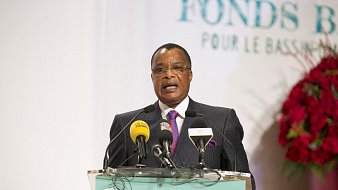 L'image montre un homme, probablement un leader ou un représentant politique, s'exprimant devant un public. Il porte un costume sombre avec une cravate rose et semble être en train de donner un discours. Devant lui, il y a un podium avec plusieurs microphones et un panneau indiquant "Fonds Bleu pour le Bassin du Congo". En arrière-plan, on peut voir des éléments décoratifs qui suggèrent un événement officiel, avec des logos et des affiches liés au thème de la conservation et du développement. L'atmosphère semble sérieuse, indiquant l'importance de l'événement.