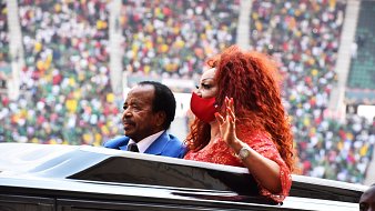 Dans cette image, nous voyons un homme et une femme qui se trouvent à l'intérieur d'une voiture décapotable. L'homme, habillé en costume, a une expression sérieuse et semble saluer la foule. La femme, aux cheveux volumineux et roux, porte une robe brillante et un masque rouge, et elle lève la main en signe de salutation. En arrière-plan, le stade est rempli de spectateurs, avec des couleurs vives de vêtements qui suggèrent un rassemblement festif. L'ambiance générale semble être celle d'un événement important, peut-être une cérémonie ou une célébration.