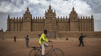 L'image montre une grande mosquée en banco, typique de l'architecture soudanaise, avec des murs en terre crue qui s'élèvent vers le ciel. Sa façade est ornée de nombreuses pointes et créneaux, formant un motif impressionnant. Au premier plan, un jeune garçon traverse la scène à bicyclette, tandis que d'autres enfants jouent et se déplacent dans l'espace. Le sol est poussiéreux, et le climat semble chaud, avec des nuages ici et là dans le ciel. L'ambiance est vivante, illustrant une scène animée de la vie quotidienne.